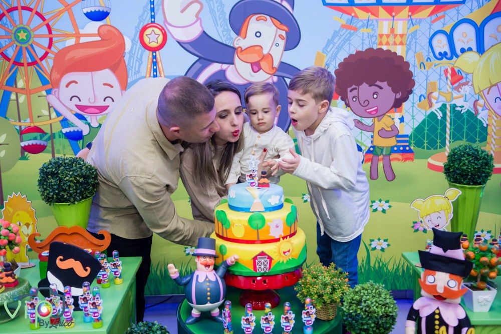 Pai, mãe e irmão soprando a vela do bolo na festa de um ano, com cenário de parque de diversões