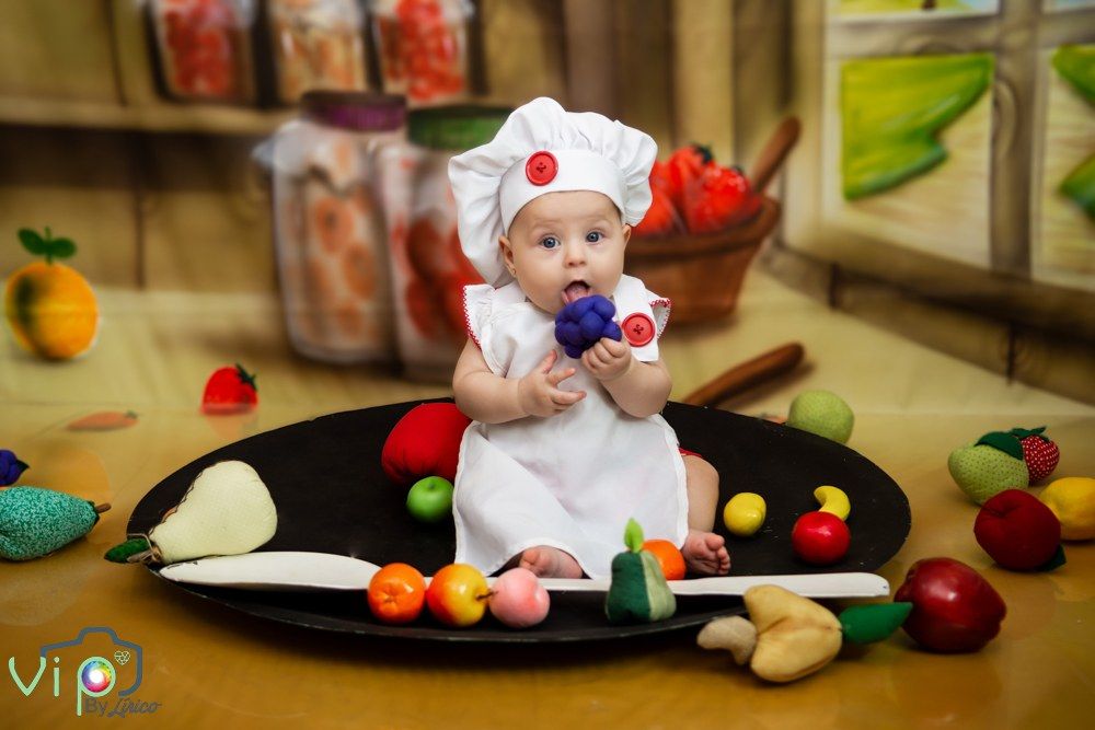 Bebê de touca de chef sentado numa frigideira de cenário, cercado de frutinhas de feltro