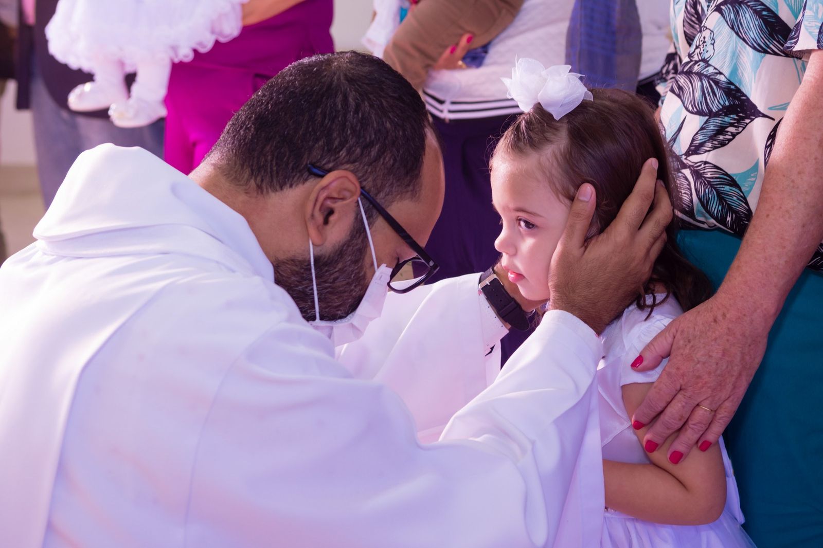 Padre com a mão sobre a cabeça da menina de vestido branco, durante o batizado