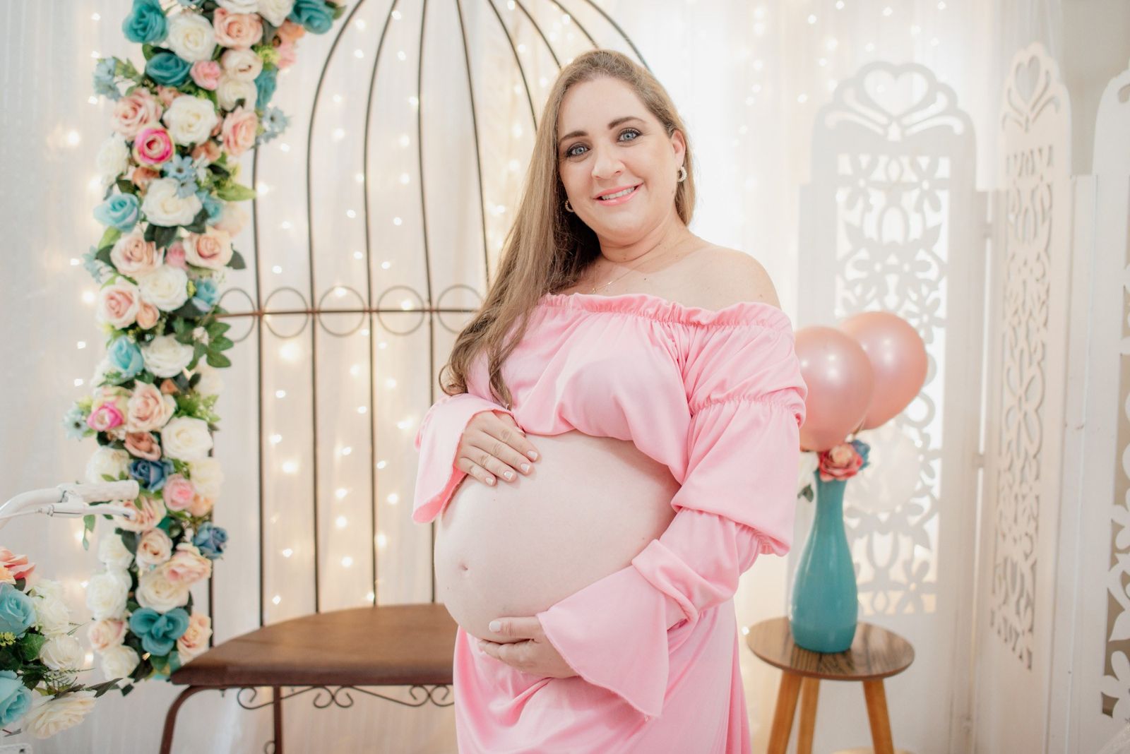 Gestante de vestido rosa, com as mãos na barriga, em cenário de flores e luzinhas no estúdio