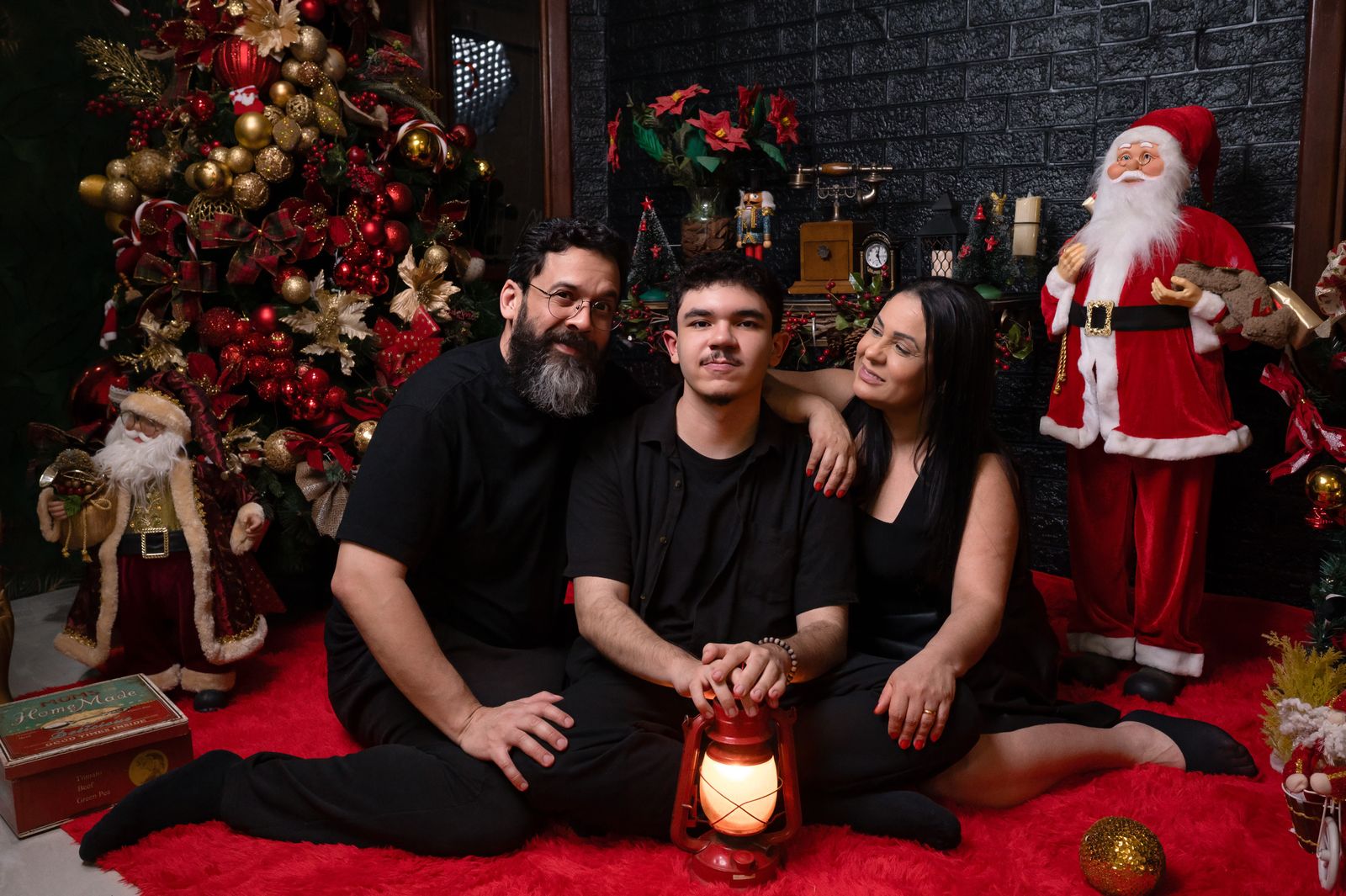 Família sentada num tapete vermelho diante da árvore de Natal, com um lampião aceso